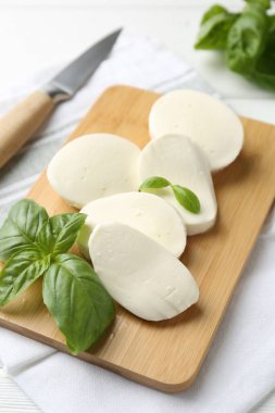 Lezzetli mozzarella peyniri parçaları, fesleğen yaprakları ve beyaz ahşap masa üzerinde bıçak, yakın plan.