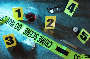 Adli tıp uzmanı. Sarı suç mahalli bandı, işaretler ve kanıtlar gri masada, üst tarafta.