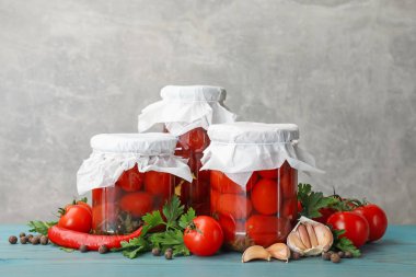 Kavanozlarda salamura domates, taze ürünler ve baharatlar açık mavi ahşap masa üzerinde gri arka planda.