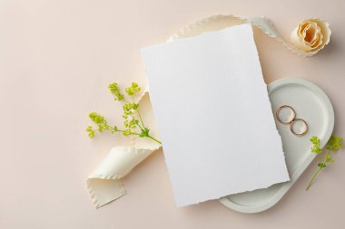 Düğün davetiyesi, altın yüzükler, çiçek dekoru ve bej arka planda kurdele. Metin için boşluk