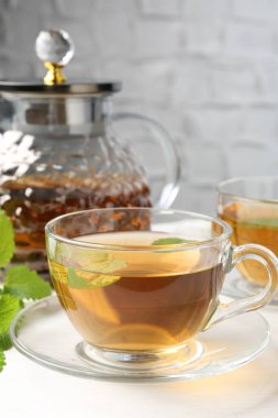 Cam fincanlarda aromatik limon merhemi çayı, çaydanlık ve beyaz masada taze yapraklar, yakın plan.