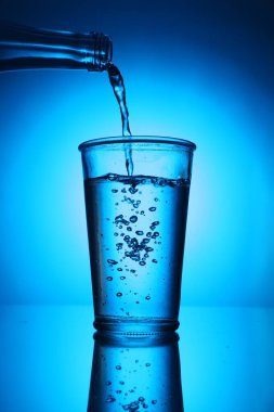 Mavi arkaplanda şişeden cama su dökmek. Arka ışıklandırma