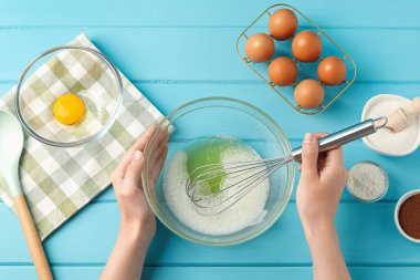 Bir kadın açık mavi ahşap masada kasede yumurta akı çırpıyor.