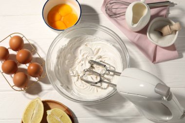Beyaz ahşap masada çırpılmış krema, yumurta ve diğer ürünlerle el çırpma makinesi.