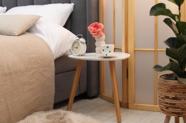 Çalar saati olan yan masa, bir fincan çay ve yatağın yanında çiçek.