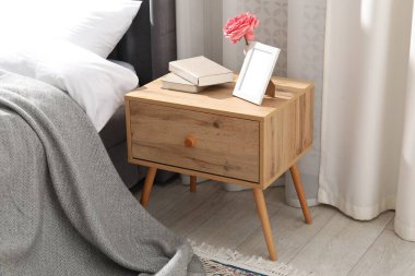 Yatağın yanında kitap, çiçek ve çerçeve olan komodin.