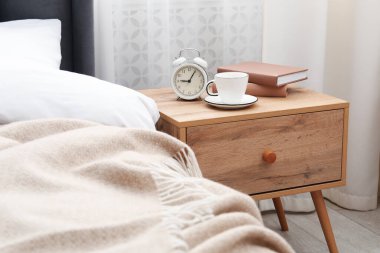 Yatağın yanında kahve, kitap ve çalar saat olan komodinin yanında.