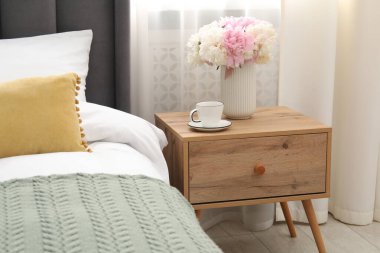 Yatağın yanında kahve ve çiçek olan bir komodin.