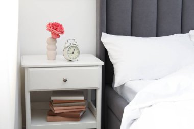 Komodinin yanında çalar saat ve yatağın yanında çiçek var.