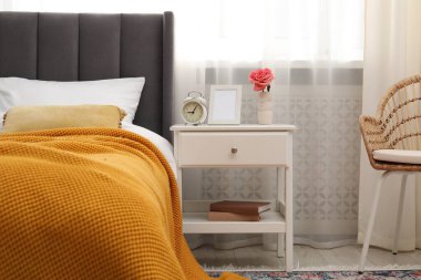 Yatağın yanında çalar saati, çiçeği ve çerçevesi olan bir komodin.