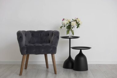 Beyaz duvarın yanında rahat gri koltuk, masalar ve vazo.