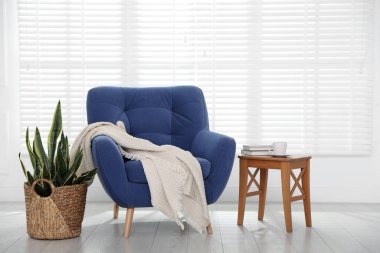 Rahat mavi koltuk, battaniye, yan masa ve beyaz duvarın yanındaki ev bitkisi.