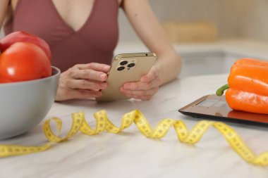 Beyaz mermer masada sebzeli akıllı telefon kullanan bir kadın, mutfak terazisi ve kapalı alanda mezura, yakın plan.