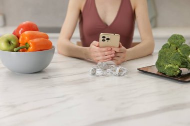 Beyaz mermer masada sebzeli akıllı telefon kullanan ve kapalı alanda şerit çeken bir kadın.