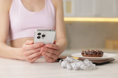 Beyaz ahşap masada akıllı telefon kullanan bir kadın. Mutfak terazisi, çörek ve kapalı alanda mezura kullanıyor.