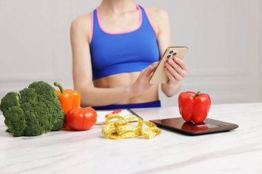 Beyaz mermer masada akıllı telefon kullanan bir kadın mutfak tartısı, sebzeler ve kapalı alanda mezura kullanıyor.