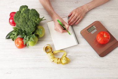 Hafif ahşap masada diyet planı geliştiren kadın mutfak tartısı, mezura ve sağlıklı yiyecekler, üst görünüm.