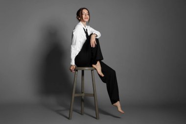 Şık giyinmiş güzel bir kadın açık gri arka planda sandalyede oturuyor.