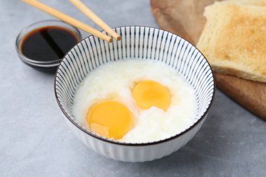Geleneksel Asya kahvaltısı kasede yarı haşlanmış yumurta, soya sosu, ekmek ve çubuklar gri masada, üst tarafta.