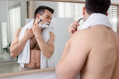 Yakışıklı adam evde aynanın yanında tıraş oluyor.