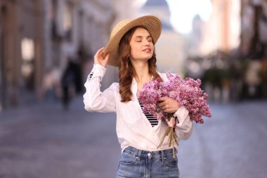 Şapkalı gülümseyen kadın, şehir caddesinde bir demet leylak çiçeğiyle.