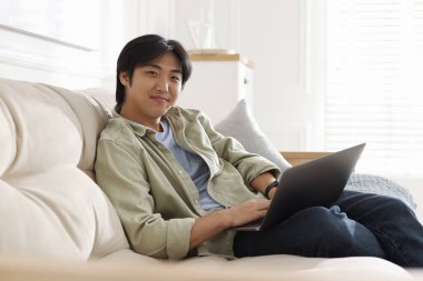 Evdeki kanepede dizüstü bilgisayarı olan yakışıklı Asyalı adam.