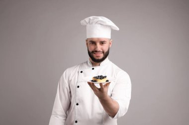 Üniformalı mutlu pastacı açık gri arka planda yaban mersinli lezzetli tart tutuyor.