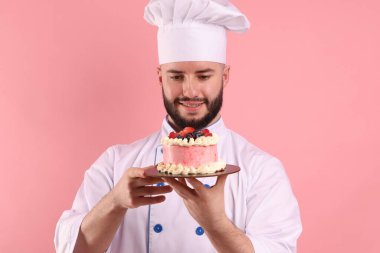 Üniformalı mutlu pastacı pembe arka planda böğürtlenli pasta tutuyor.