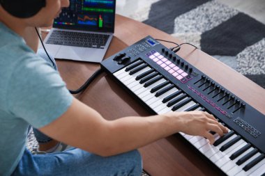 MIDI klavyesi ve dizüstü bilgisayarıyla çalışan adam kapalı bir masada, yakın plan.