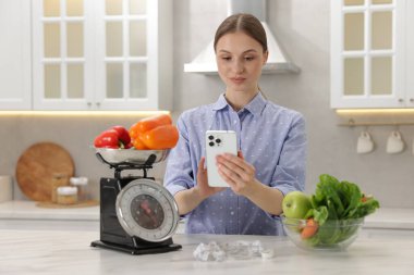 Beyaz mermer masanın yanında akıllı telefon kullanan bir kadın. Mutfak terazisi, sağlıklı yiyecekler ve kapalı alanda mezura kullanıyor.