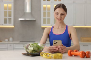 Beyaz masada akıllı telefon kullanan bir kadın mutfak tartısı, salata, mezura ve dambıl kullanıyor.