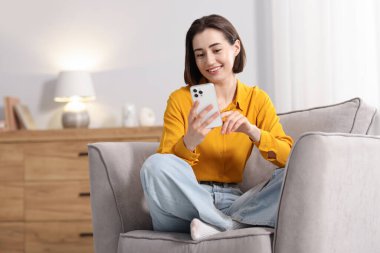 Evdeki koltukta modern akıllı telefon kullanan bir kadın.