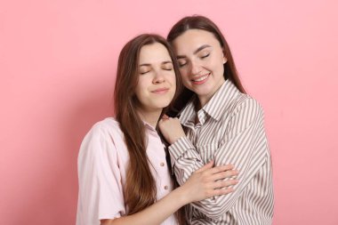 Arkadaşlar pembe arka planda birbirlerine sarılıyorlar.