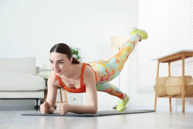 Spor kıyafetli genç bir kadın evde kalas egzersizi yapıyor.