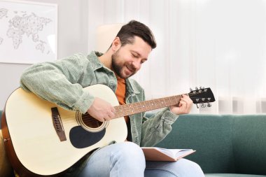 Rahatlatıcı hobi. Evdeki kanepede gitar ve kitapla gülümseyen adam.