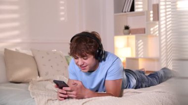 Kulaklıkla gülümseyen genç çocuk akıllı telefon kullanıyor ve evde yatakta müziğin keyfini çıkarıyor.