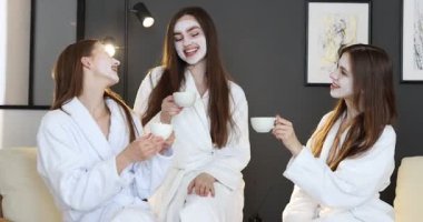 Yüzü maskeli mutlu kadınlar spa ameliyatından sonra bornozlu sağlıklı içecekler içerler.