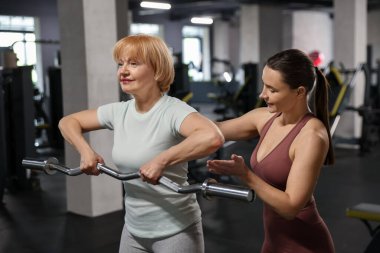 Kıdemli kadın spor salonunda profesyonel antrenör tarafından yönetilen EZ kıvrım çubuğuyla egzersiz yapıyor.