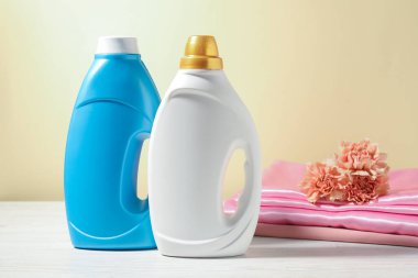 Kumaş yumuşatıcı şişeler, temiz kıyafetler ve karanfil çiçekleri açık sarı arka planda beyaz ahşap masa üzerinde