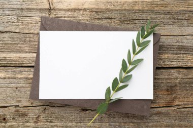 Boş davetiye kartı, zarf ve yeşil dal, ahşap masa, üst manzara.