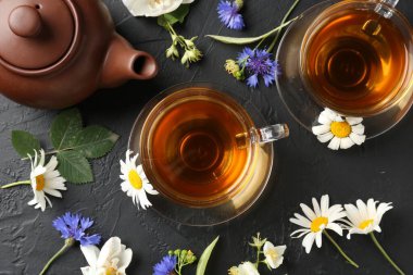Cam fincanlarda aromatik bitki çayı, farklı çiçekler ve siyah masada çaydanlık.