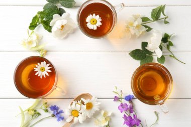 Cam fincanlarda aromatik bitki çayı, farklı çiçekler ve beyaz ahşap masada yeşil yapraklar, düz yatak. Metin için boşluk