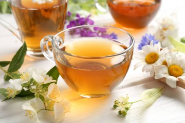 Cam bardaklarda romatik bitki çayı ve beyaz ahşap masada farklı çiçekler, yakın plan.