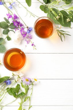 Cam fincanlarda aromatik bitki çayı, farklı çiçekler ve beyaz ahşap masada yeşil yapraklar, düz yatak. Metin için boşluk