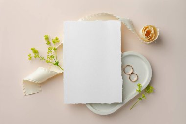 Düğün davetiyesi, altın yüzükler, çiçek dekoru ve bej arka planda kurdele.