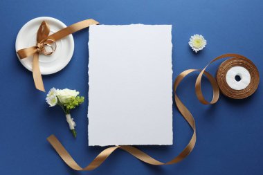 Düğün davetiyesi, altın yüzükler, çiçek dekoru ve mavi arka planda kurdele.