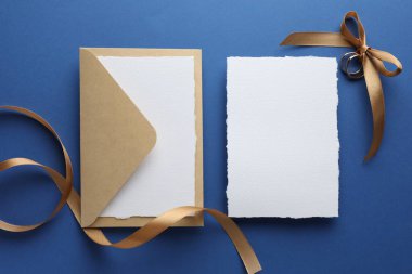 Düğün davetiyeleri, zarf, altın yüzükler ve mavi arka planda kurdele, düz yatış