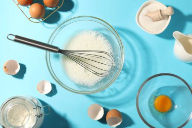 Fırça çırpma, yumurta beyazı kasede ve diğer ürünler açık mavi masada, düz yerde.