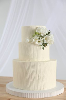 Ahşap masadaki çiçek desenli nefis düğün pastası ve beyaz arka plandaki perdeler.