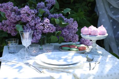 Güzel leylak çiçekleri, tatlılar, tabaklar, çatal bıçak takımı, bardaklar ve bahçede yanan mumlar.